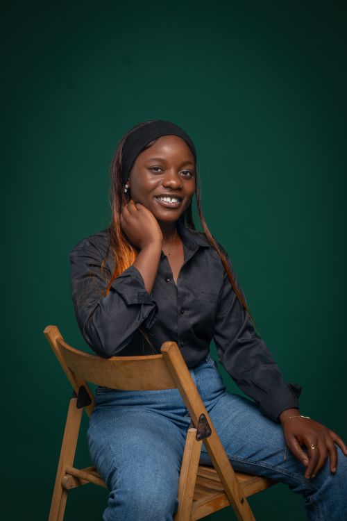 Free Smiling woman in casual attire sitting on a wooden chair against a green background. Stock Photo