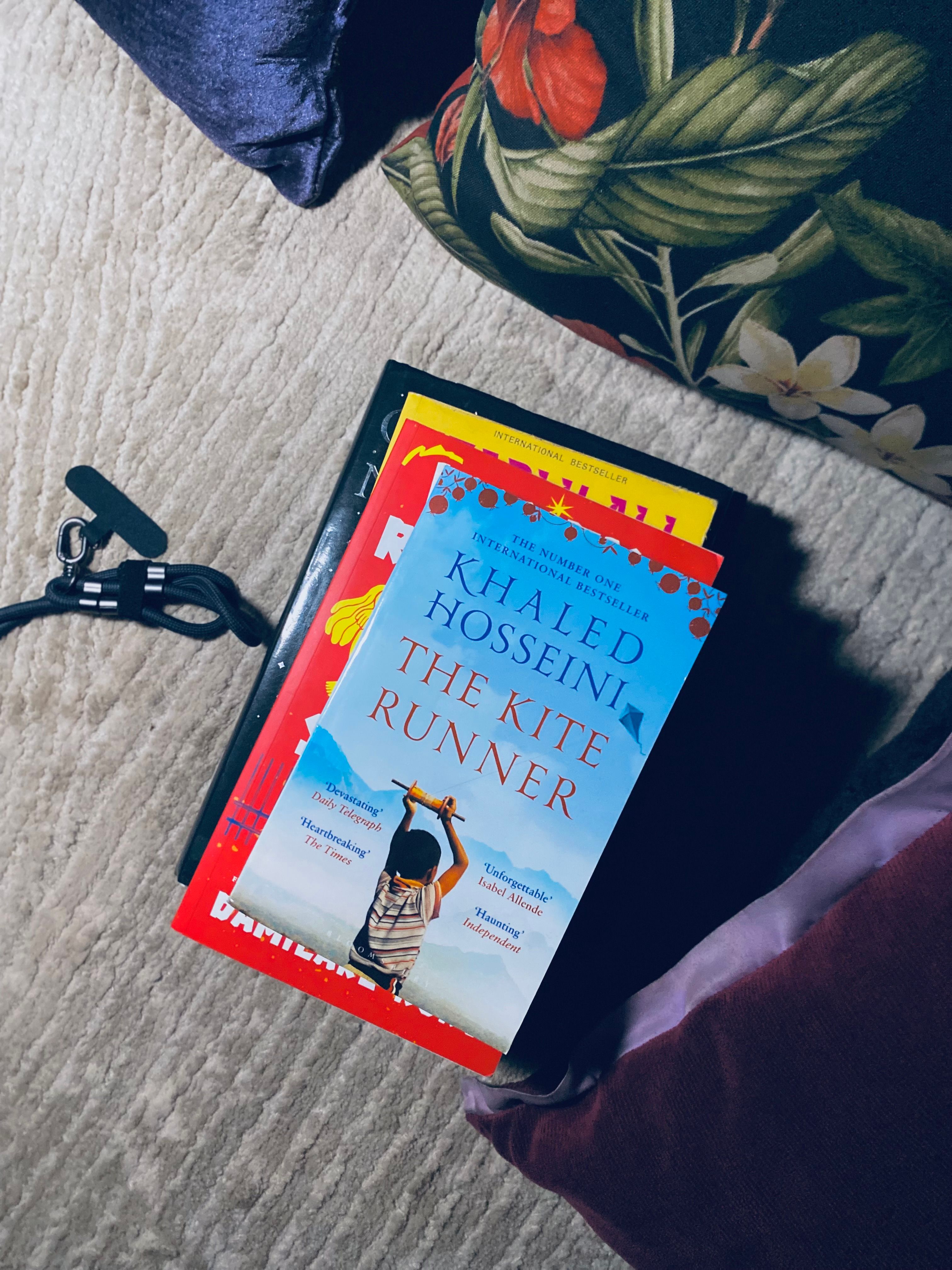 A top-down view of books, including 'The Kite Runner,' on a carpeted floor with a clip and pillow.