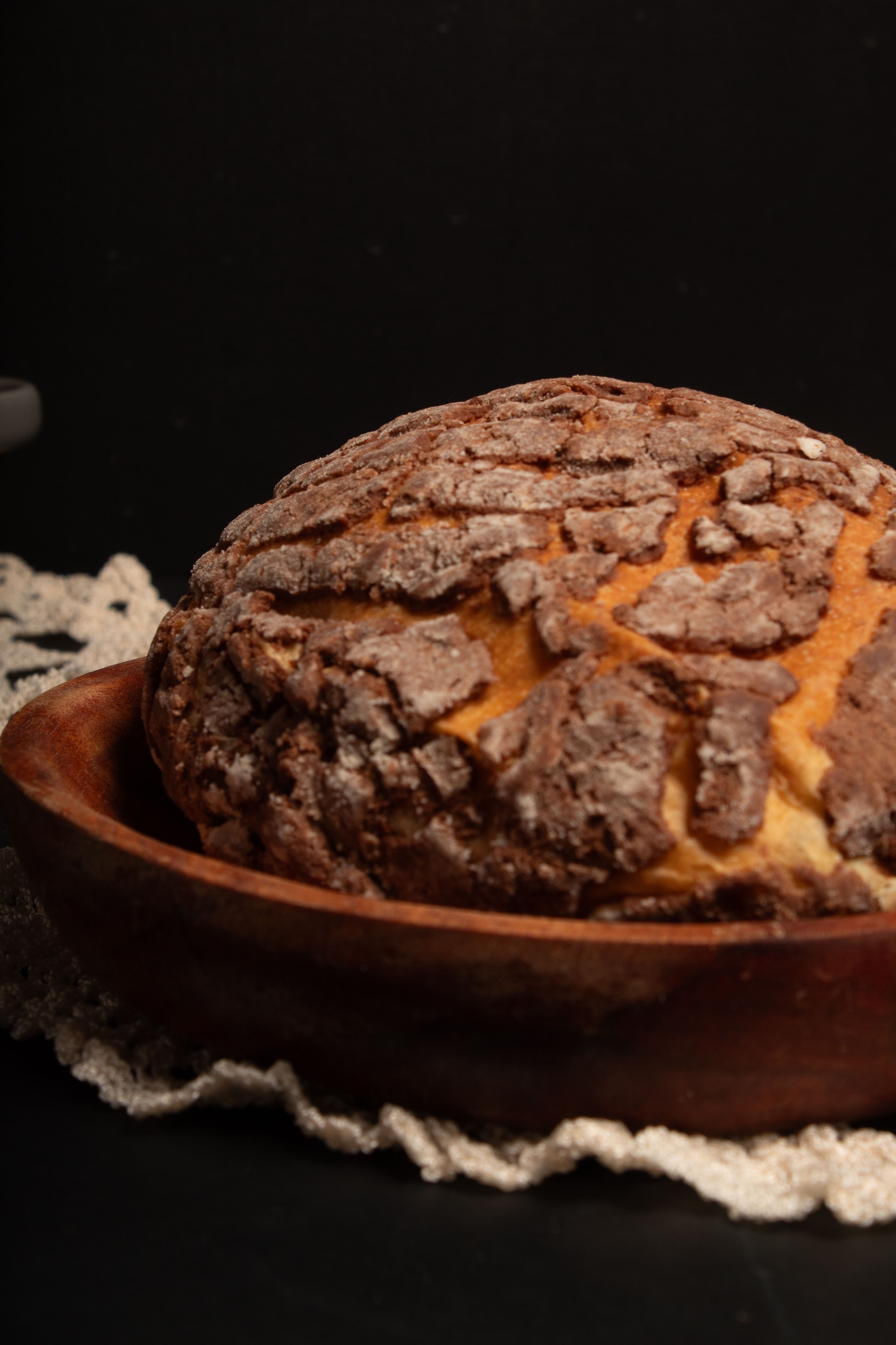 A beautifully baked concha bread sits in a rustic wooden bowl against a black background.
