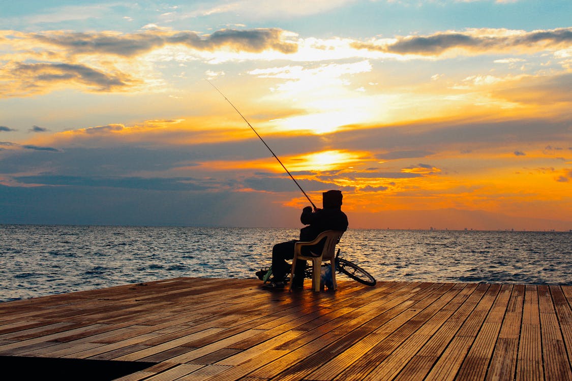 Free Silhouette of Person Sitting on Chair Holding Fishing Rod Near Body of Water during Golden Hour Stock Photo
