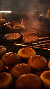 Grilled burgers and toasted buns sizzling on a barbecue, surrounded by fire and smoke.