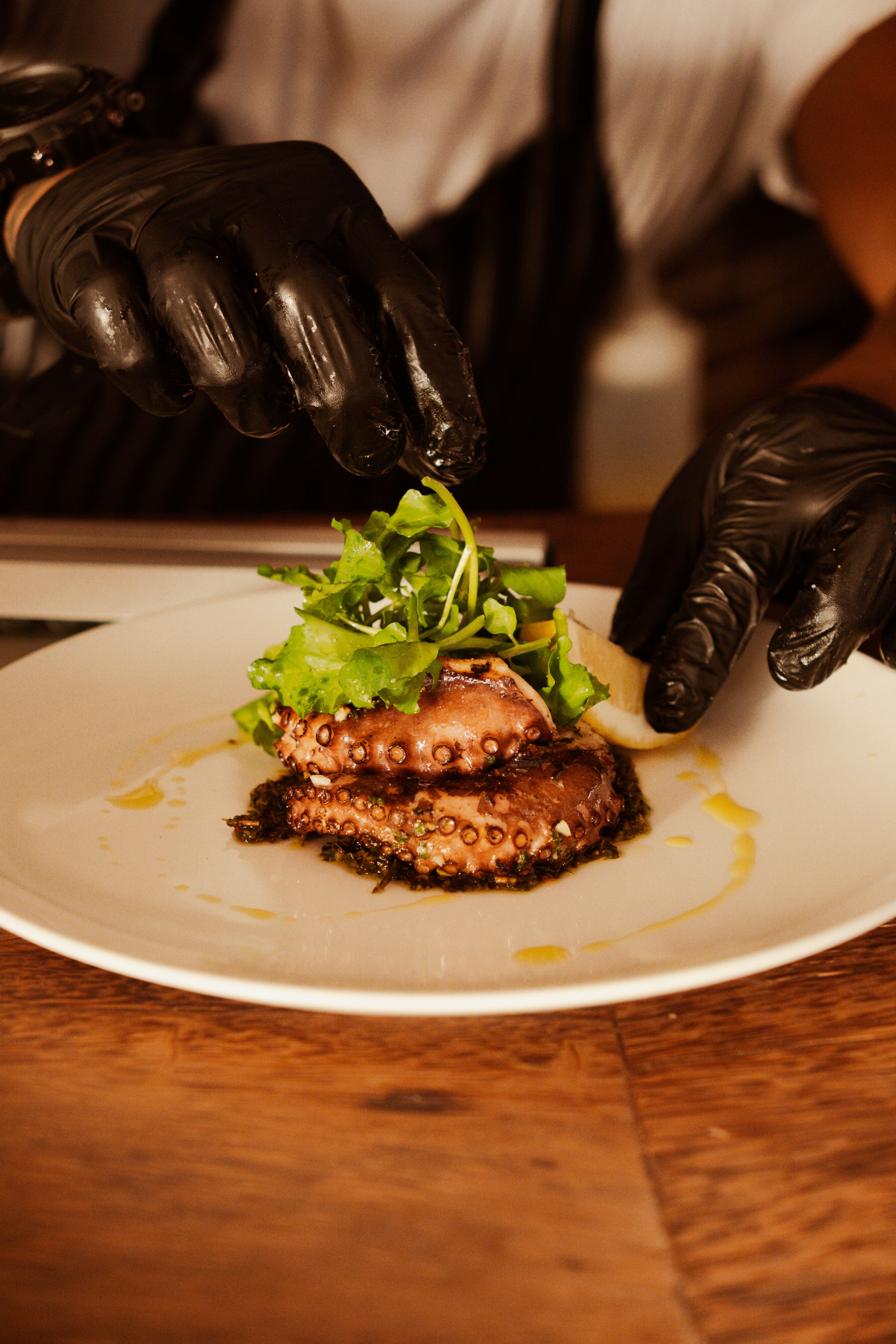 Mains gantées de noir dressant deux tranches de poulpe grillé et de la salade sur une assiette blanche