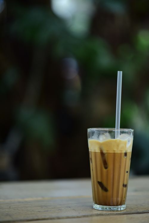 Free Glass of iced coffee with a straw on a wooden table, surrounded by greenery. Stock Photo