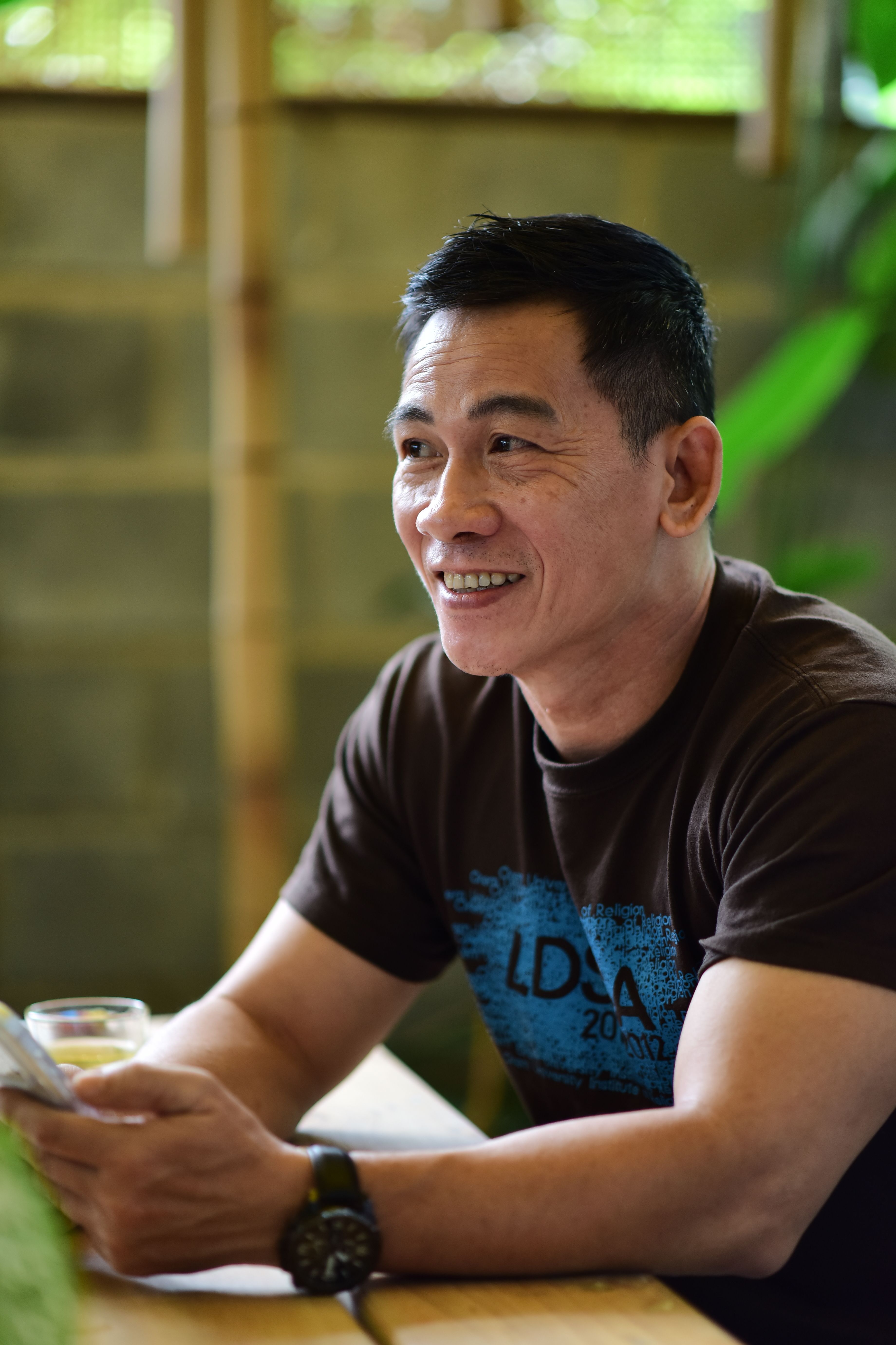 Asian man sitting in cafe with a smile, holding a smartphone.