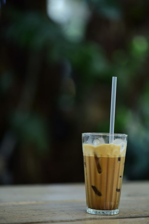 Free A glass of iced coffee with a straw on a wooden table outdoors against a blurred background. Stock Photo