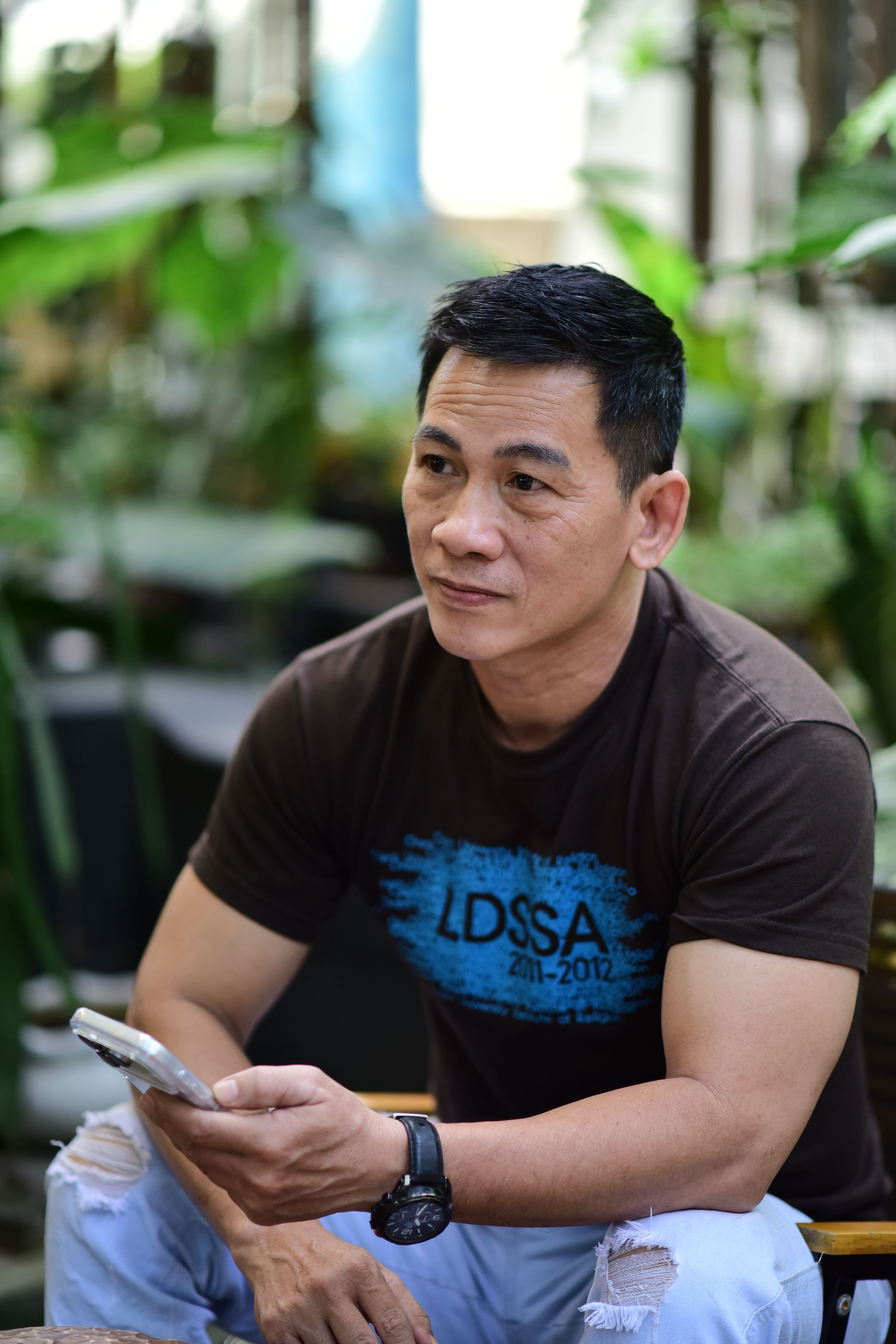 Man in casual attire holding smartphone, outdoors, surrounded by greenery.