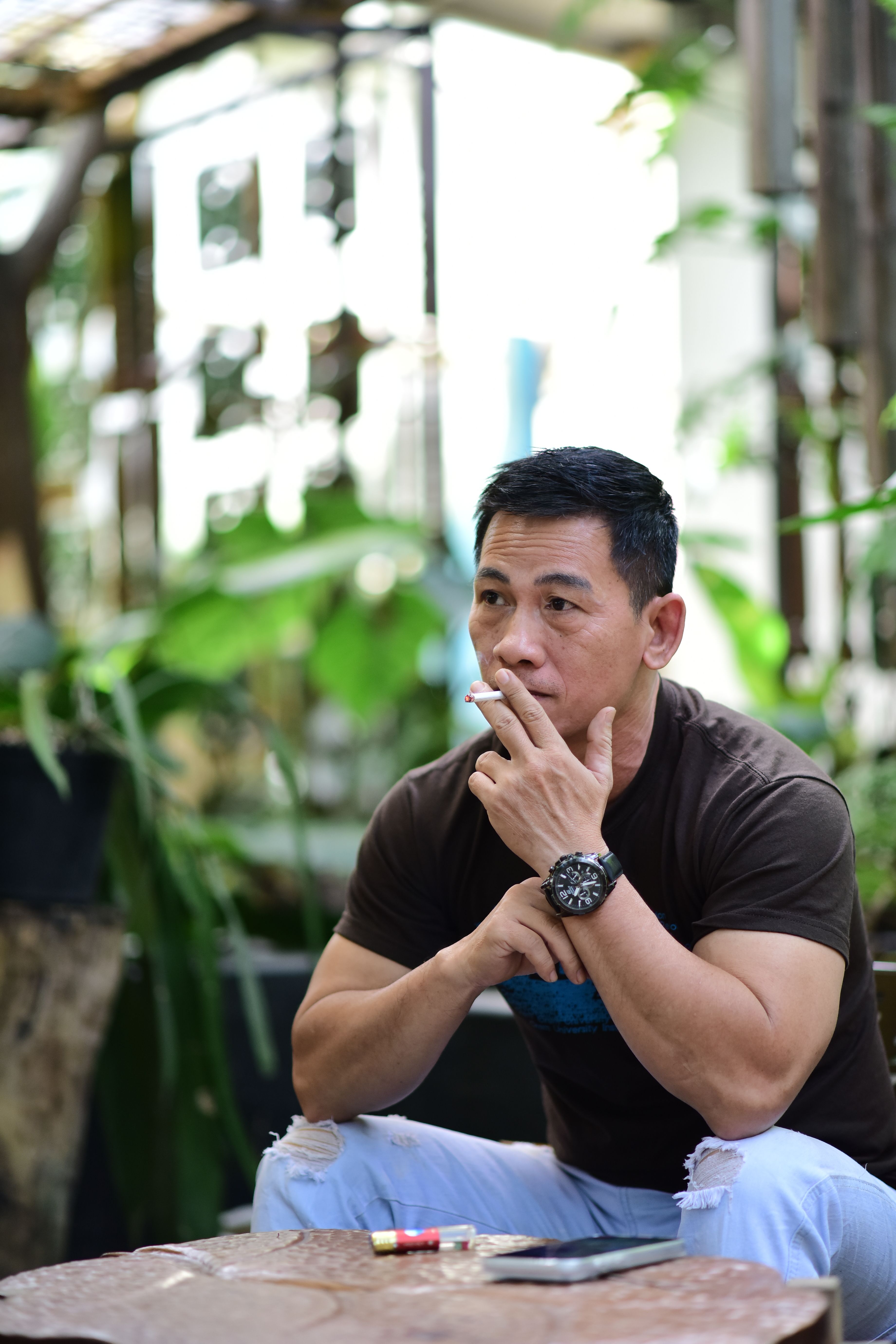 Man in casual attire sitting pensively indoors with green plants around.