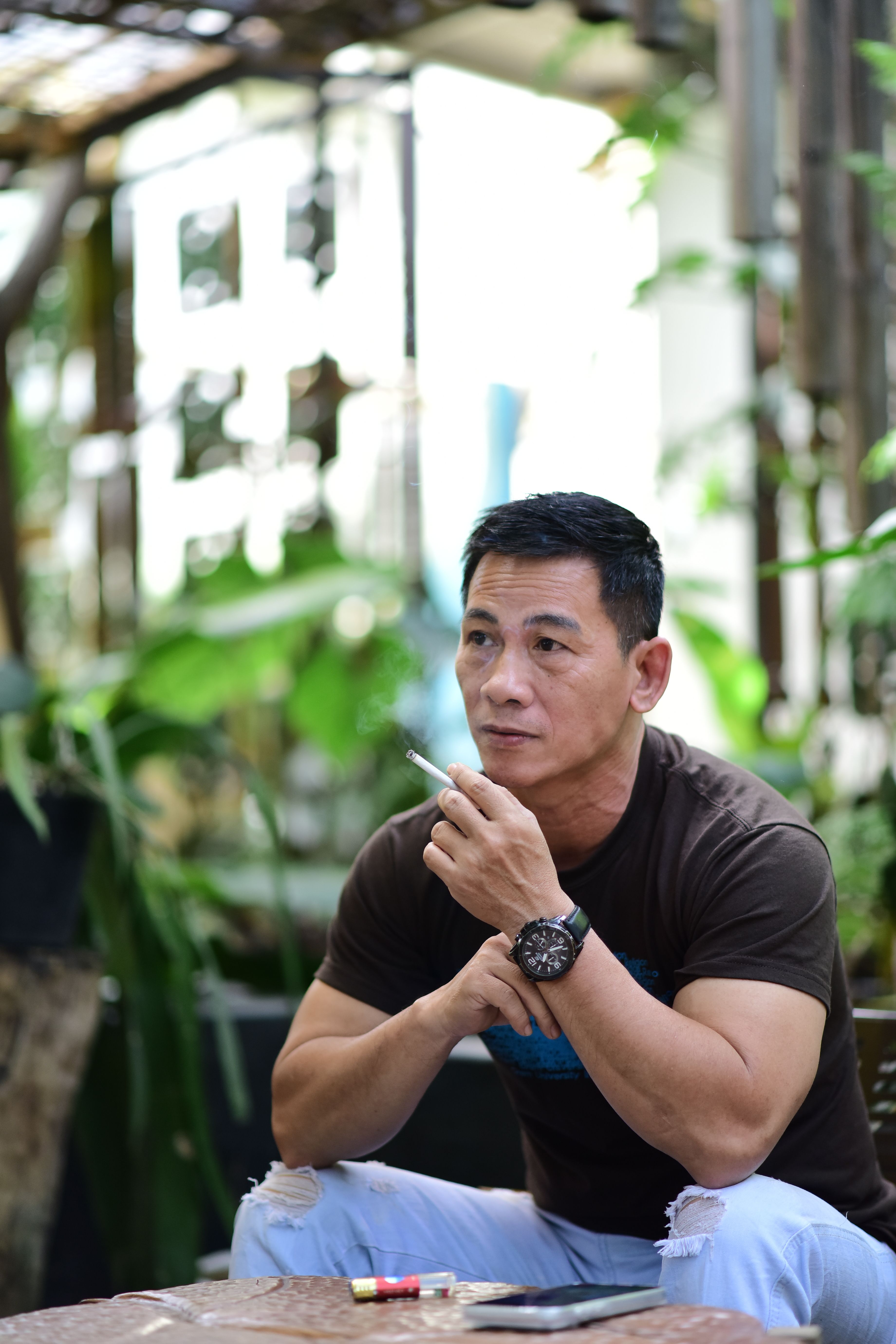 Man sitting outdoors, appearing thoughtful, surrounded by greenery.