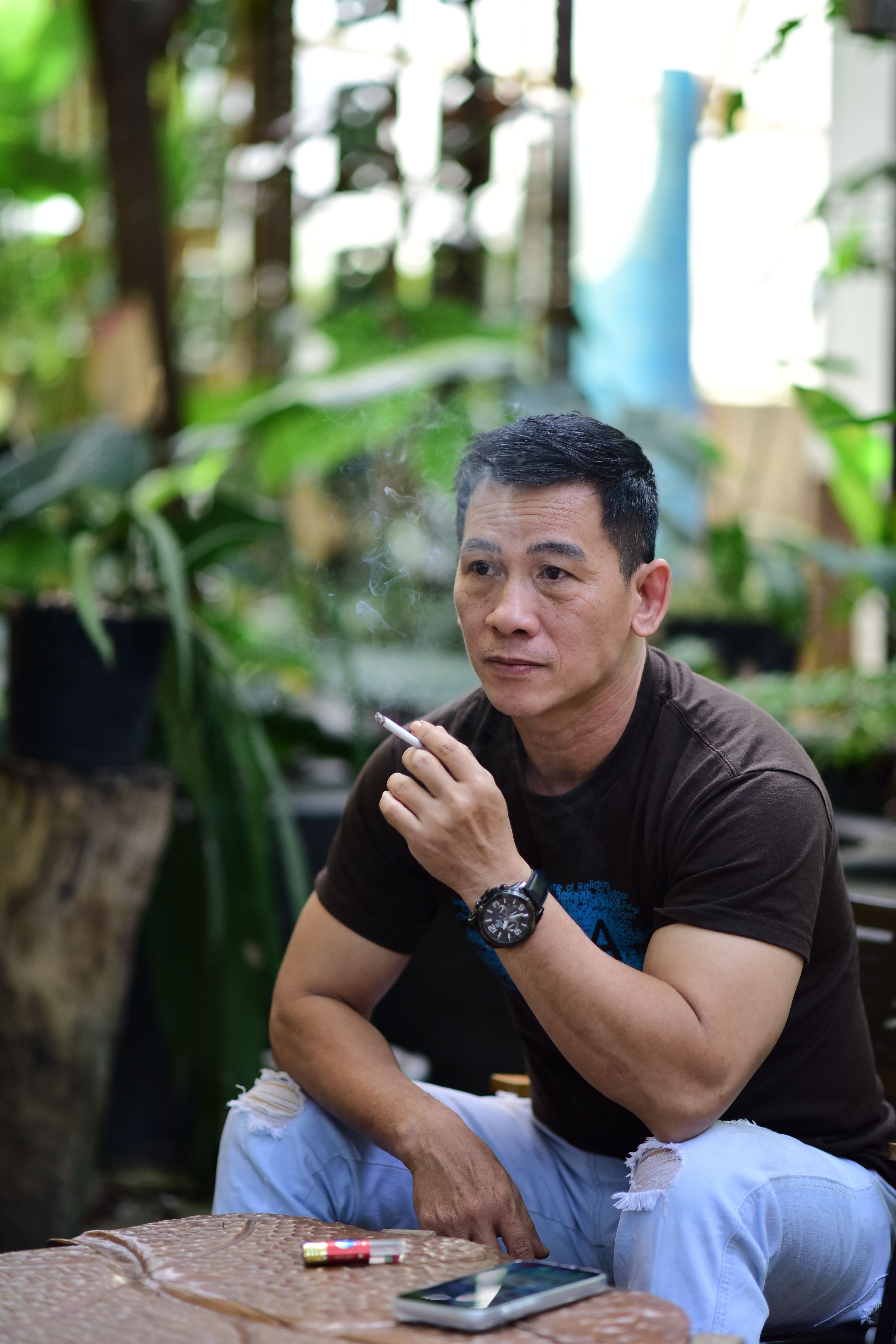 Adult man sitting outdoors, smoking, with a serene garden backdrop.