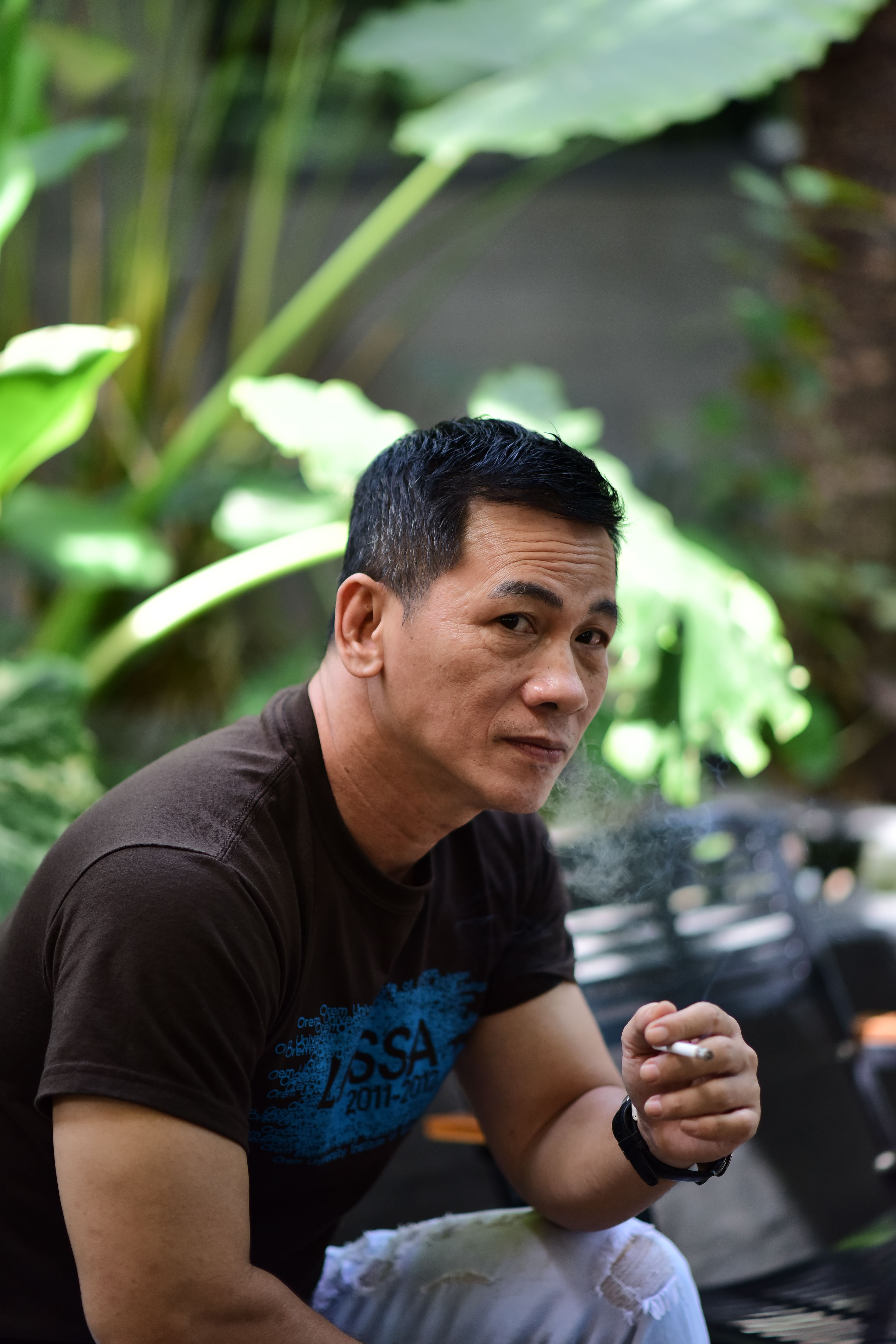 A man in a brown shirt casually smoking outdoors with lush greenery in the background.