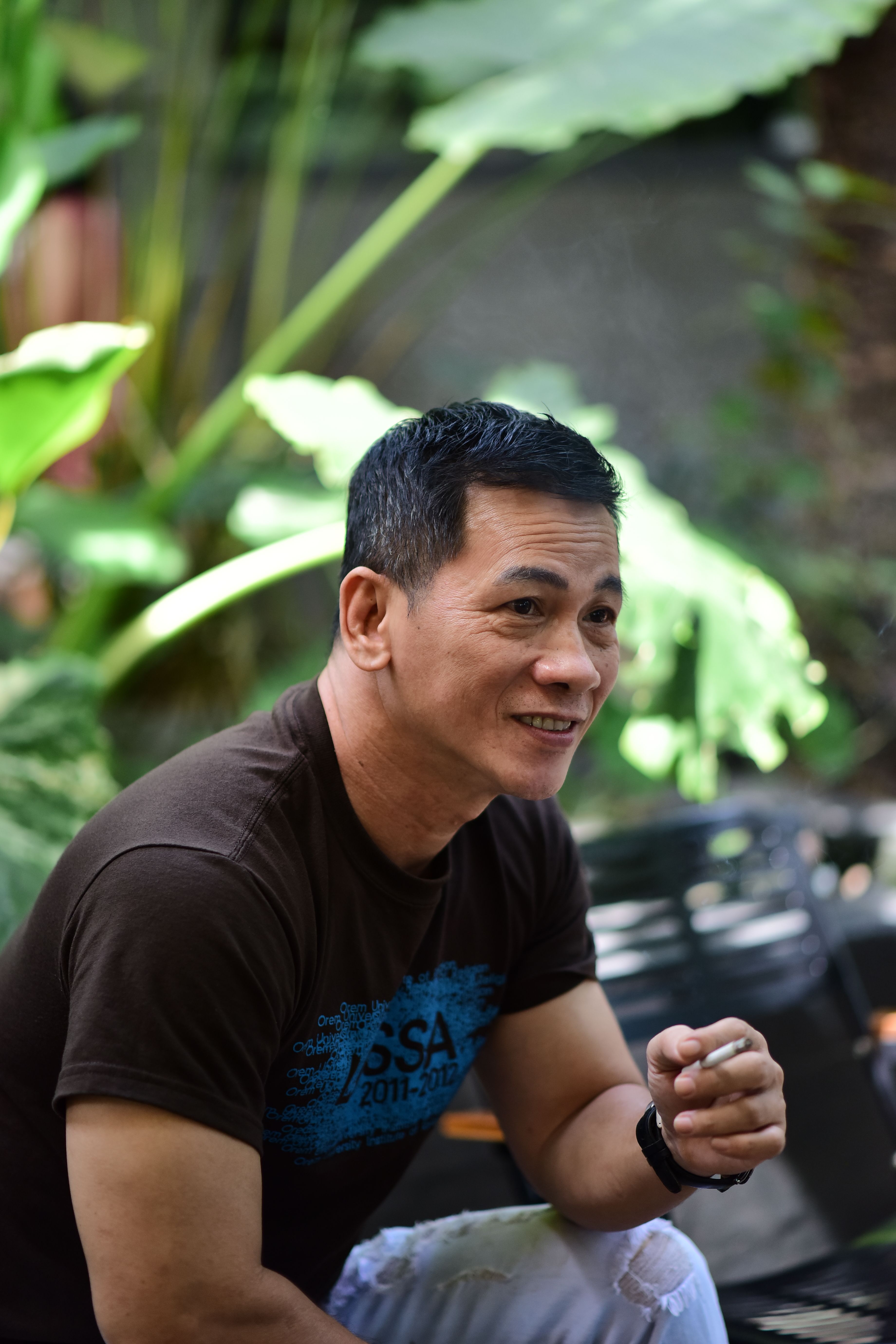 A cheerful man smiling while sitting outdoors in a lush garden environment.