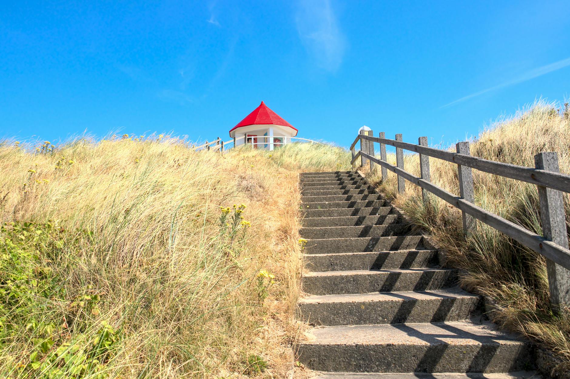 Charming pathway up sandy dune stairs to a gazebo with a red roof under a bright blue sky.