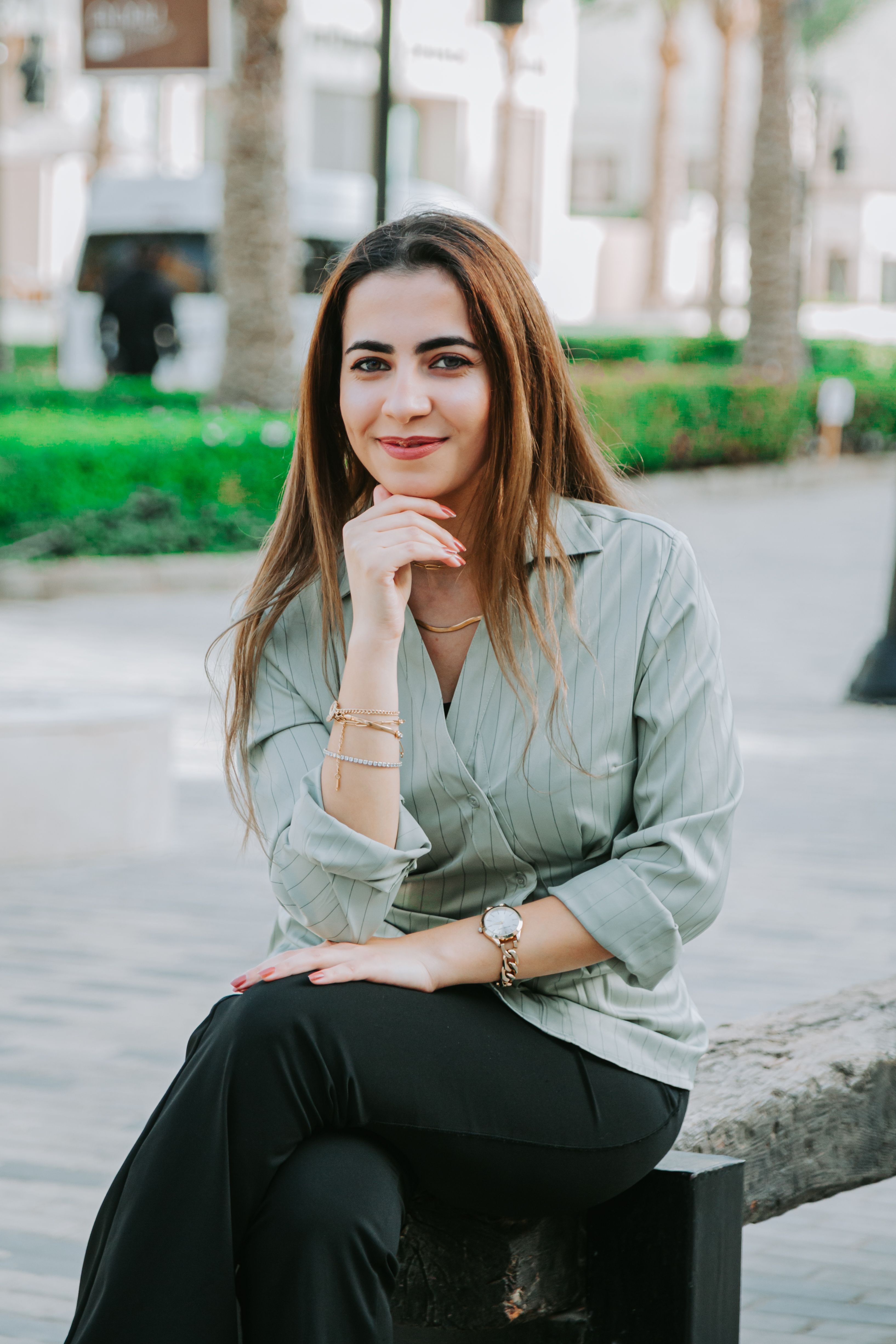 Confident young woman sitting on a bench outdoors, smiling in stylish attire, enjoying a pleasant day.