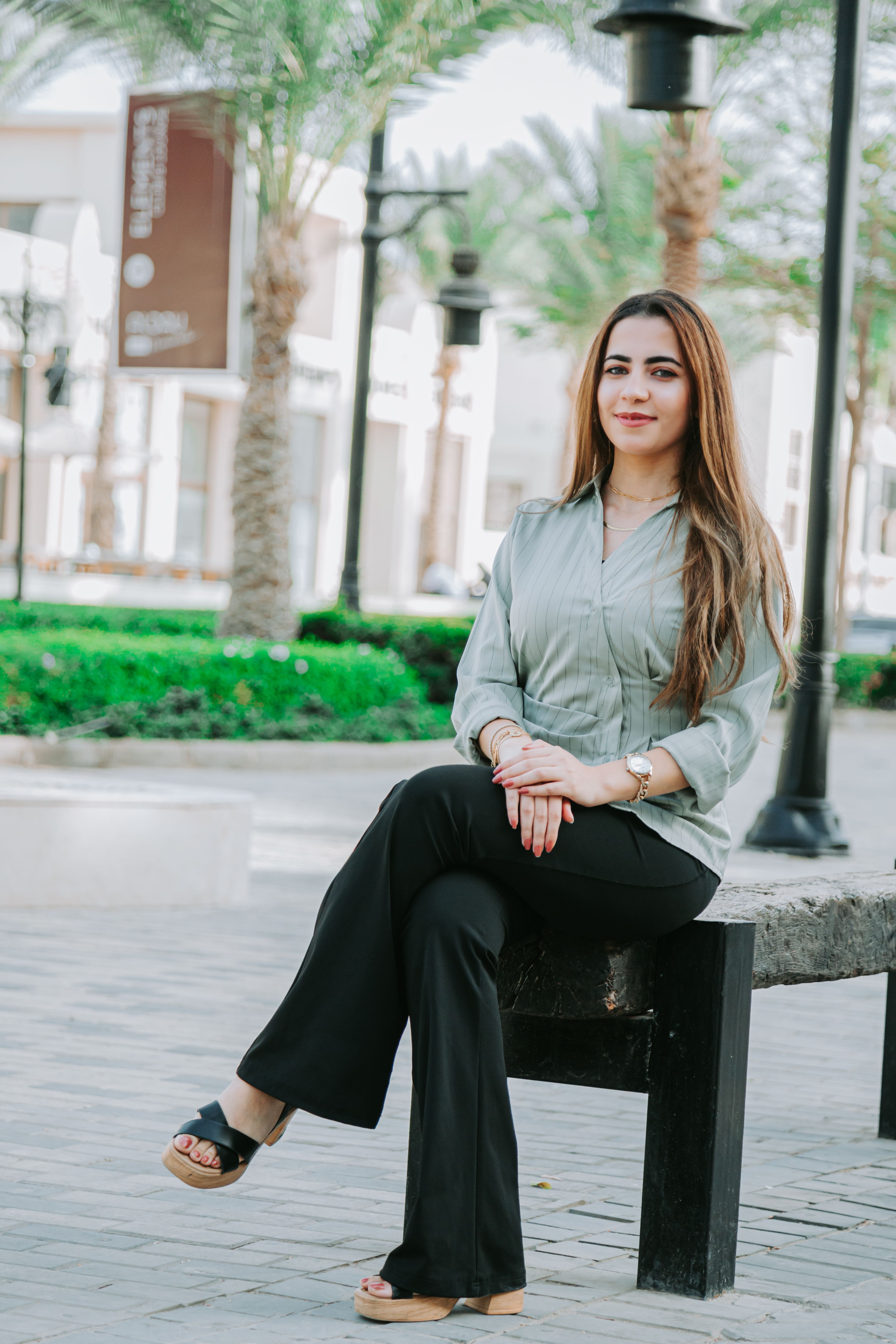 Confident woman in casual attire sitting on a bench in a modern urban setting, surrounded by palm trees.