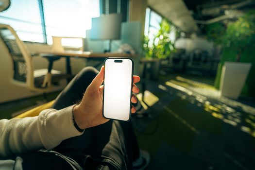A hand holds a smartphone with a blank screen in a modern, plant-filled office setting.