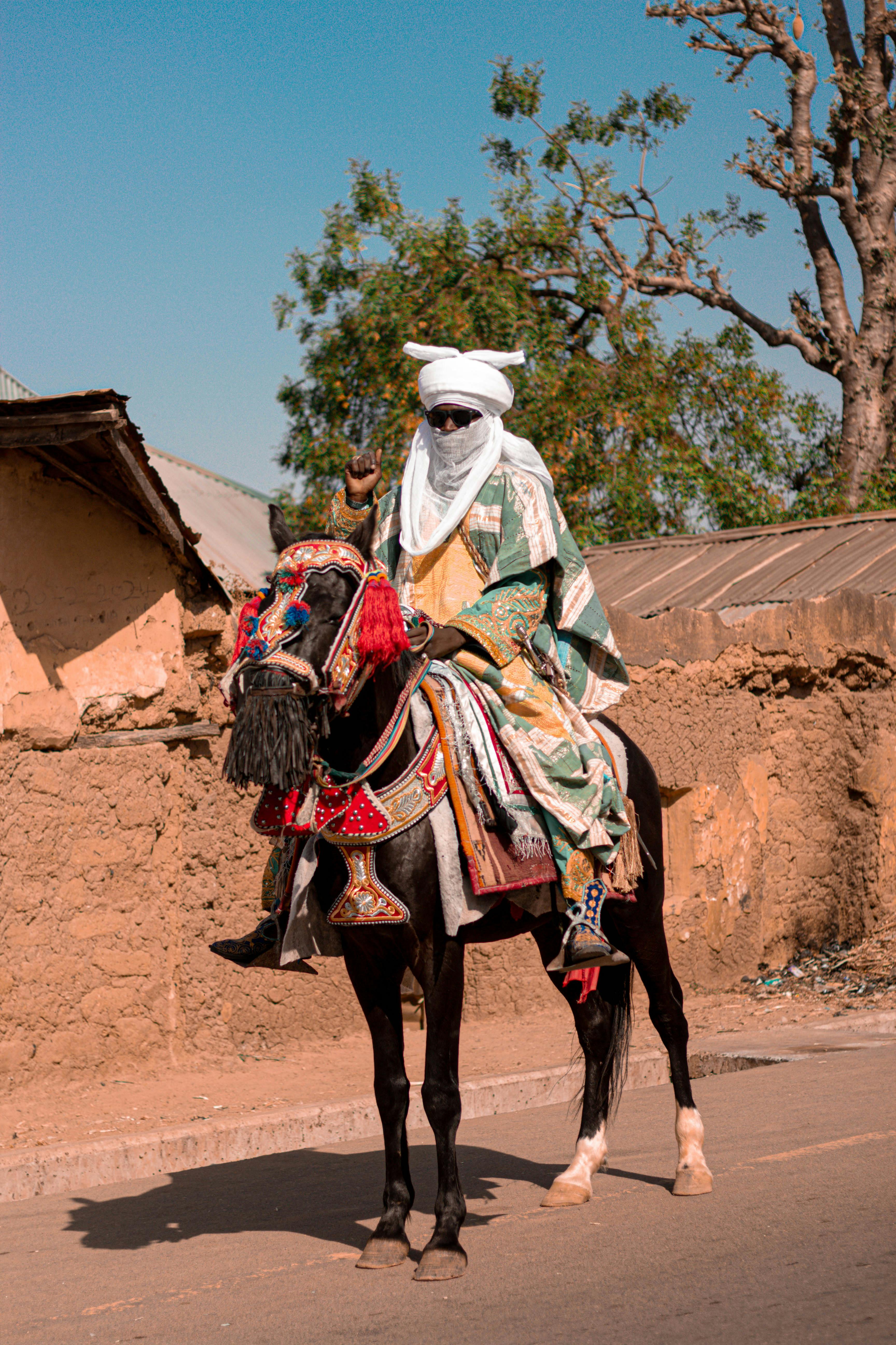 A traditional horseman in colorful attire riding on a decorative horse in a rustic setting.