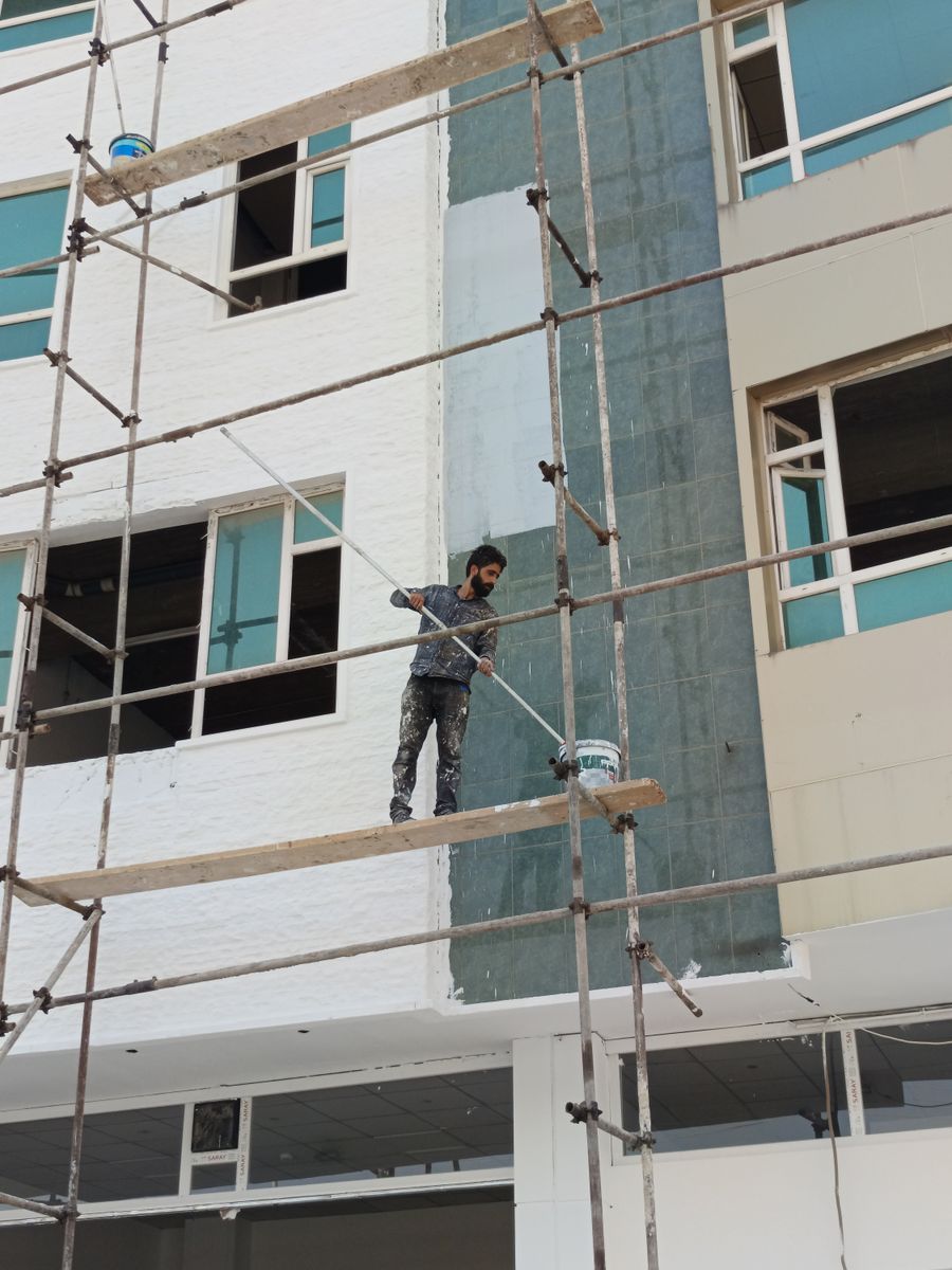 Worker standing on metal scaffolding rolling paint onto the upper exterior wall of a teal building with white window frames