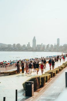 People strolling along the bustling Benidorm beach promenade on a sunny day with city skyline.