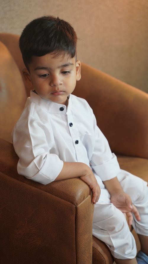 Free Young child in traditional white attire seated indoors on a brown chair, looking thoughtful. Stock Photo