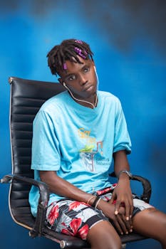 Stylish young man with earphones, sitting casually on a chair against a blue background.