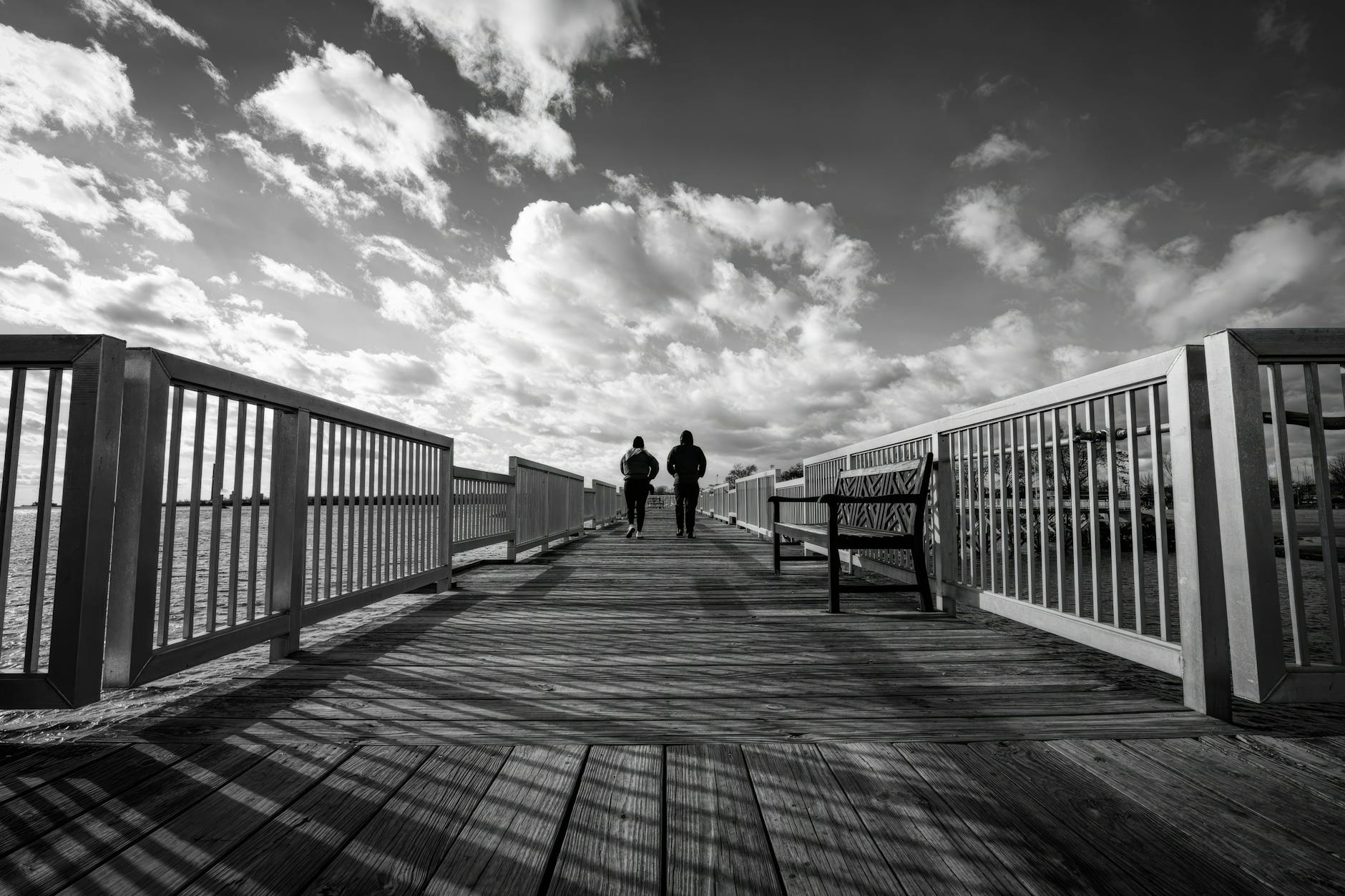 Two people walking on a pier under a dramatic cloudy sky. Captures a moody, contemplative scene.