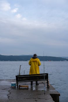 Fisherman in a yellow raincoat standing near the Bosphorus in a serene, cloudy setting.