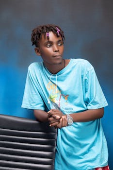 A young African man in a blue t-shirt poses casually with a relaxed expression against a gradient background.