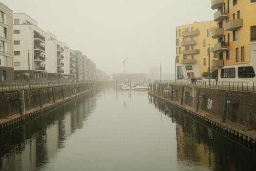 A misty canal lined with modern apartment blocks on a serene day, reflecting urban architecture.
