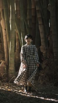 A woman in a plaid dress walking elegantly through a bamboo forest with sunlight filtering through.