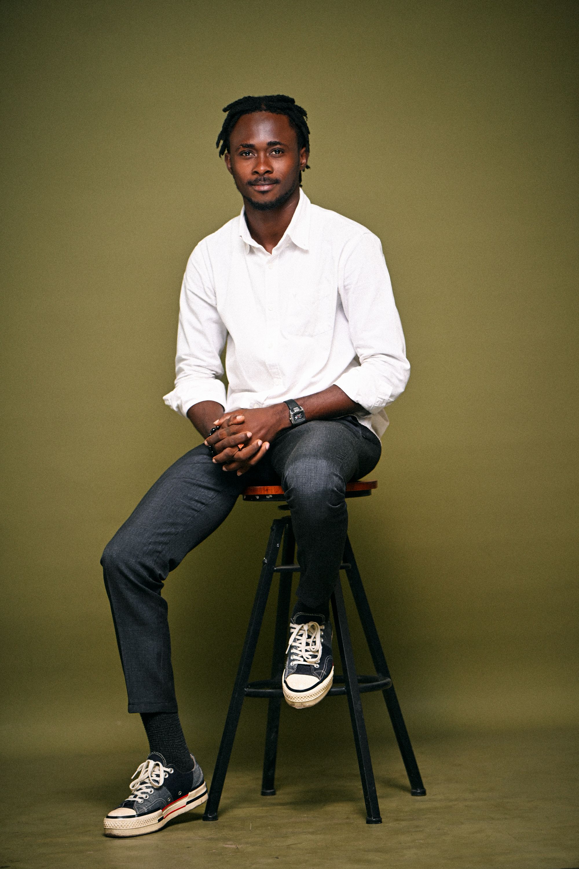 Confident young man in casual attire sitting on stool against green background.