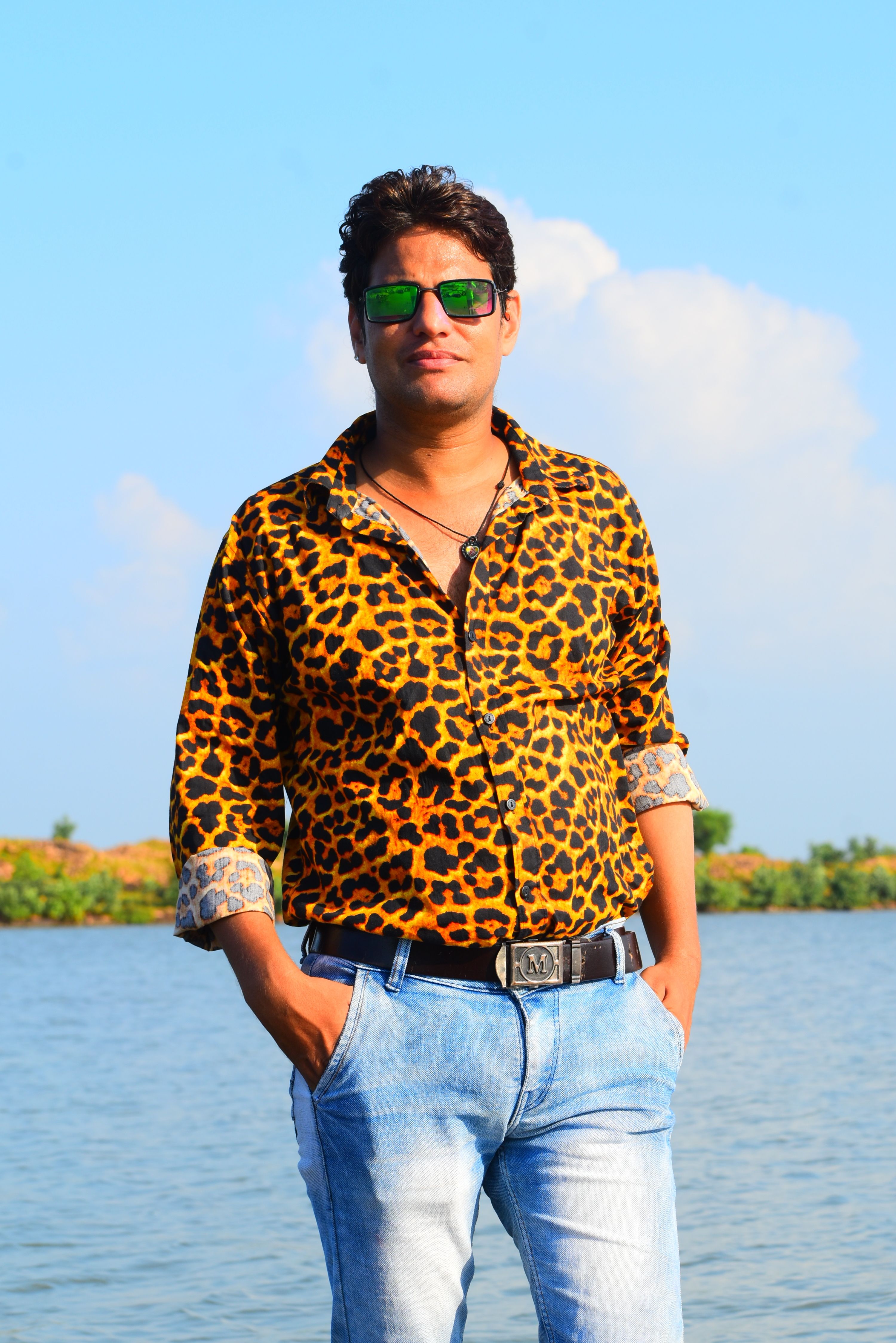A man in sunglasses and leopard print shirt stands by a scenic water backdrop.