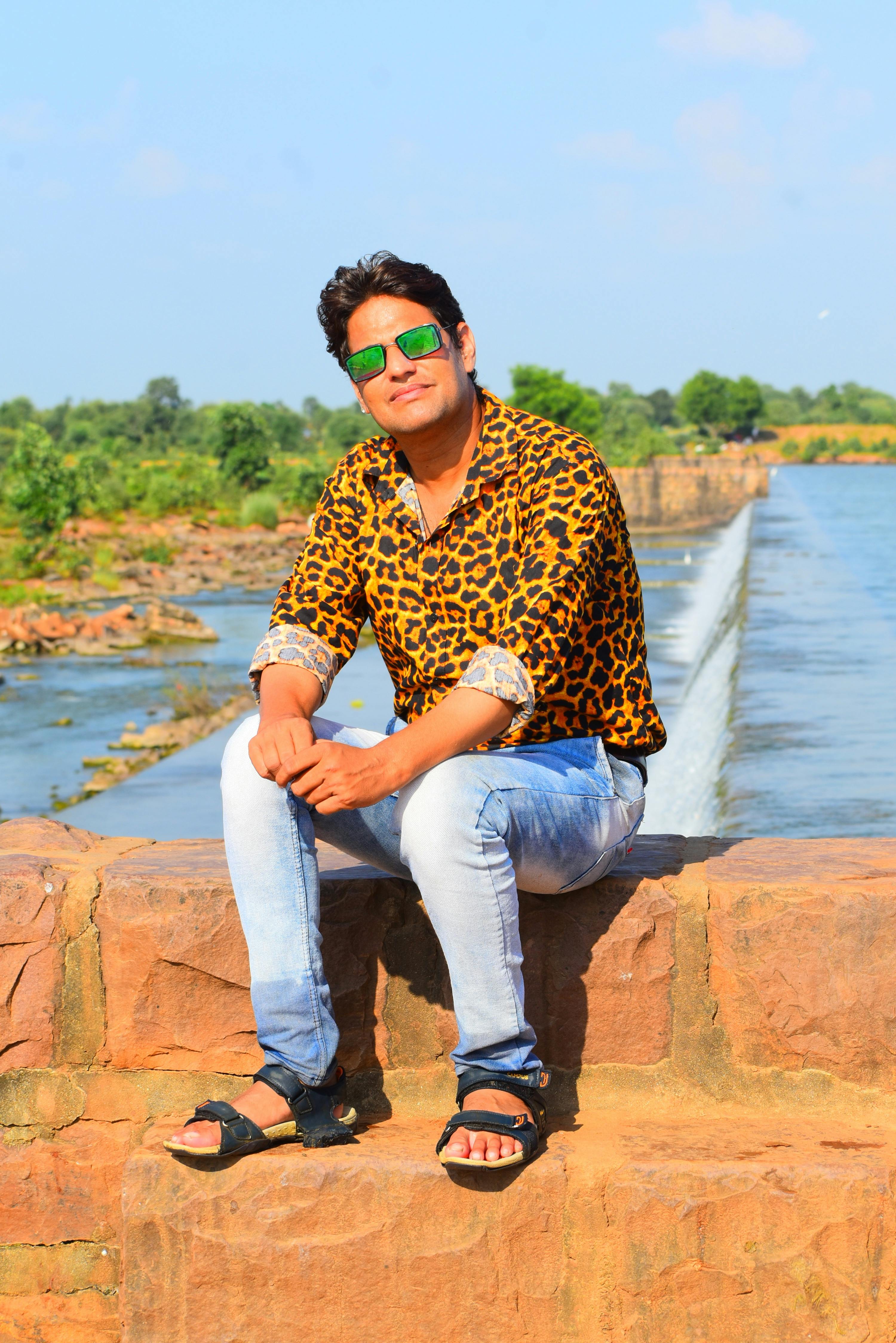Casually dressed man sitting by a river outdoors on a sunny day, wearing sunglasses and a leopard print shirt.