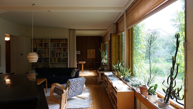 Spacious living room with large windows, natural light, and indoor plants creating a warm atmosphere.