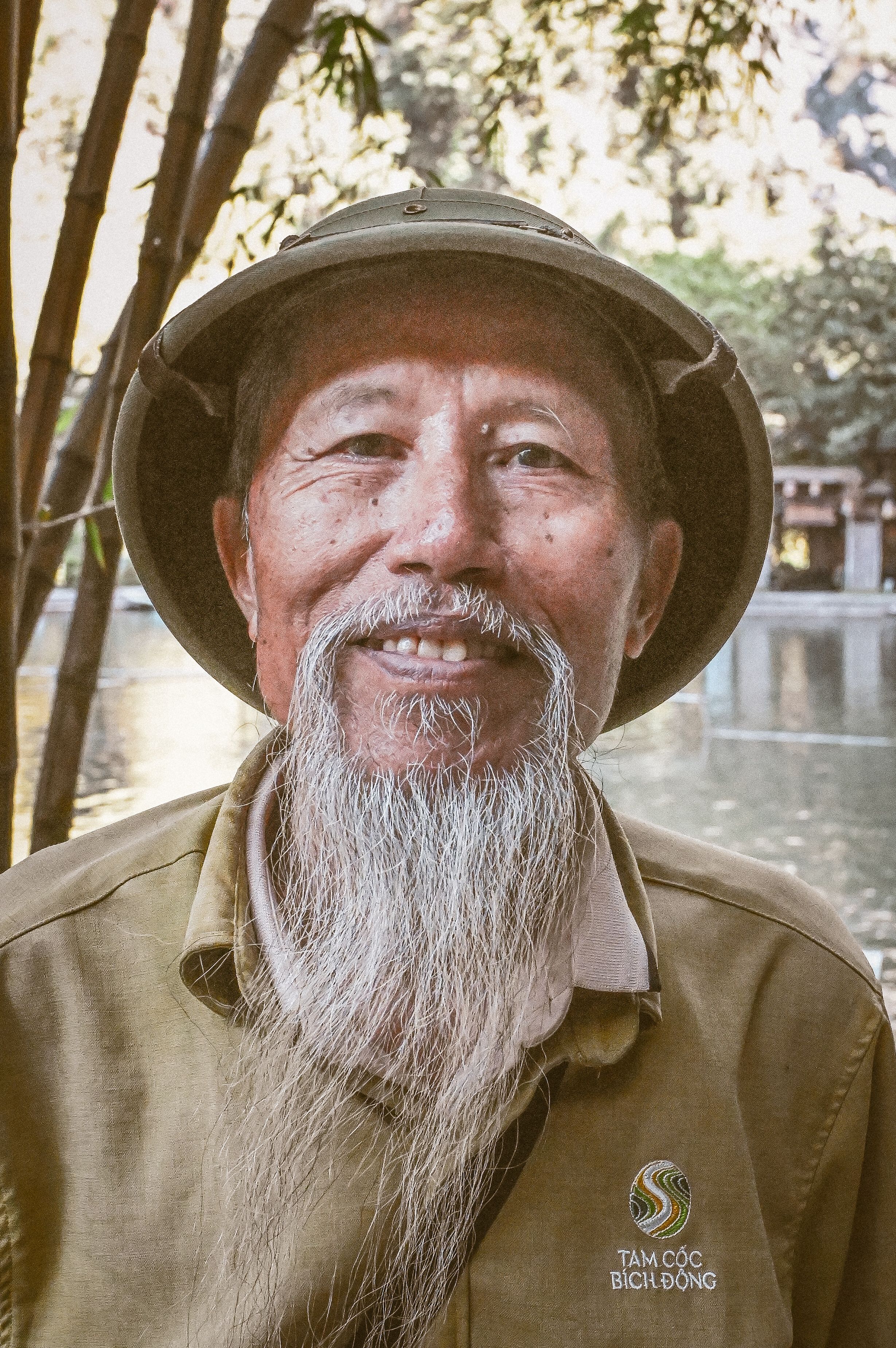 Portrait of an elderly man with a long beard and hat, smiling outdoors near water, surrounded by nature.