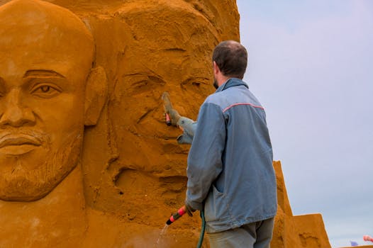 Artist meticulously works on a detailed sand sculpture outdoors, showcasing artistic skill and creativity.