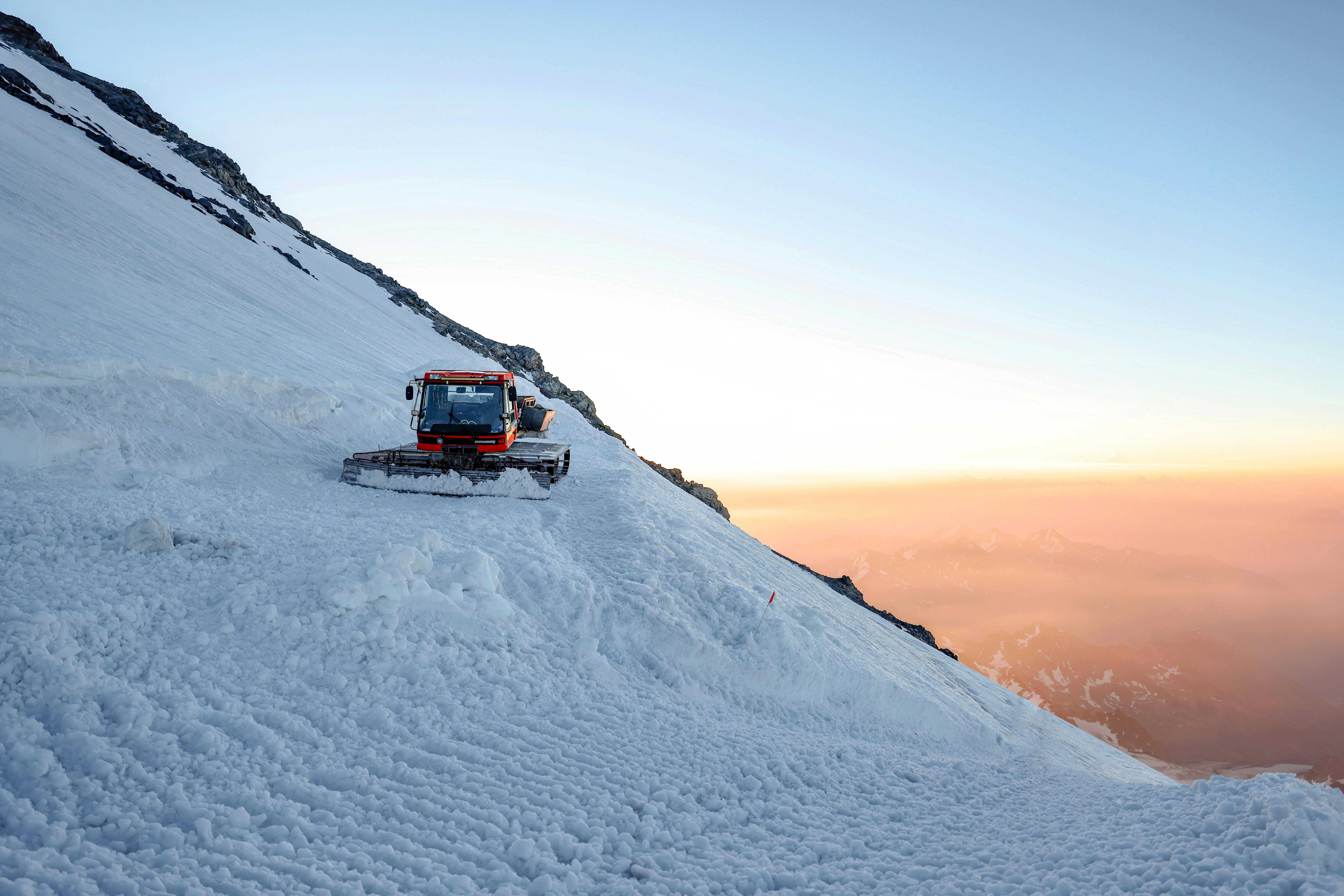 A snow groomer traversing a snowy Alpine mountain slope during a stunning sunset.