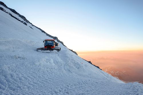 Free A snow groomer traversing a snowy Alpine mountain slope during a stunning sunset. Stock Photo