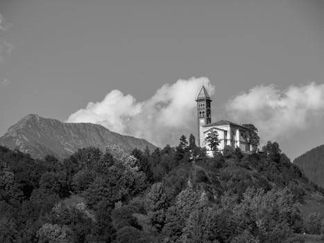 A dramatic black and white photo of a church on a forested hilltop with mountains behind.