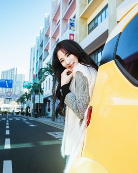 Smiling young woman in cozy sweater leaning against yellow car on bright city street.