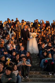 Wedding couple stands joyfully amidst a diverse crowd on steps at sunset.