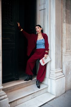Stylish woman in red suit holding a newspaper, posing confidently in an elegant urban setting.