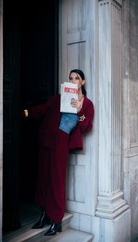 Fashionable woman in red suit with newspaper at marble entrance.