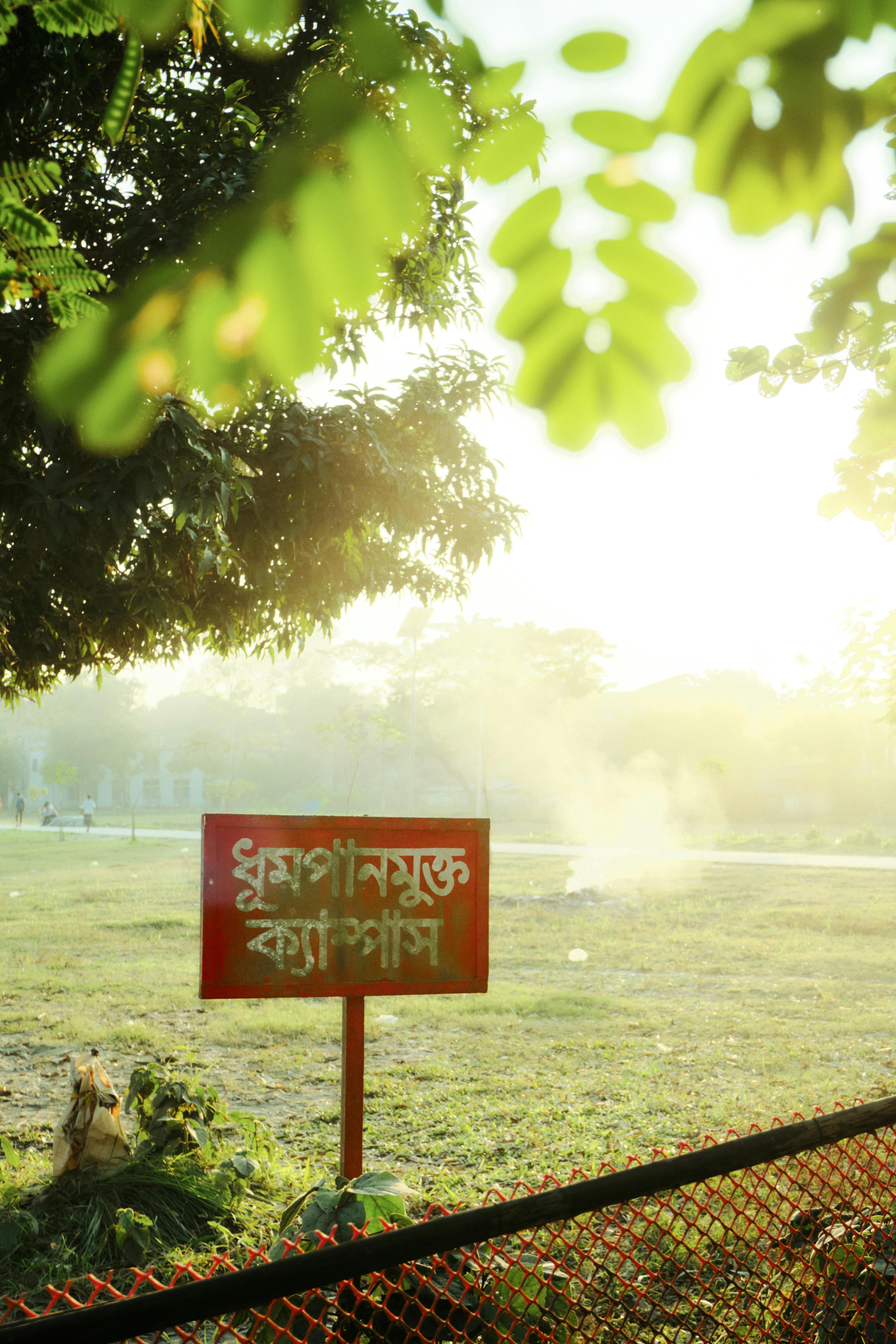 A sign in Bangla indicating a no-smoking zone in a sunny park area of Kurigram.