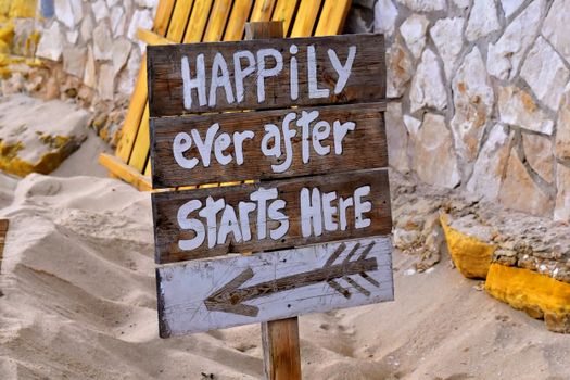 Rustic wooden sign with 'Happily Ever After Starts Here' at a sandy beach in Salema, Portugal.