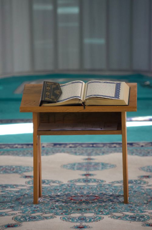 Free Open Quran resting on a stand in a serene mosque interior with ornate carpets. Stock Photo