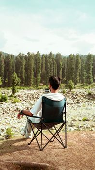 A man relaxes on a chair facing a scenic forest and rocky terrain in Kashmir, India.