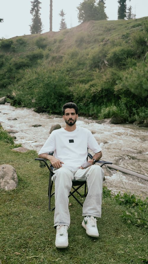 Free Man sitting on a chair by a serene river in the lush green landscape of Kashmir. Stock Photo