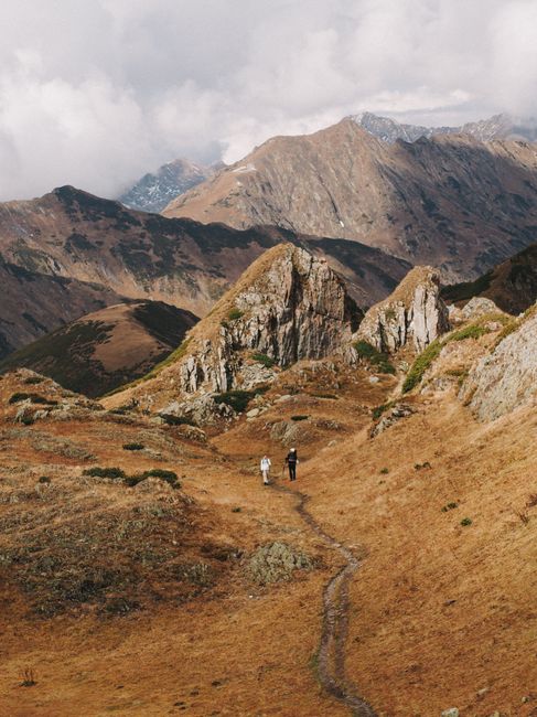 Two hikers traverse a stunning mountain trail under cloudy skies.