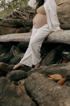 A pregnant woman in white clothing sits on a log by the rocky seaside, capturing serenity.