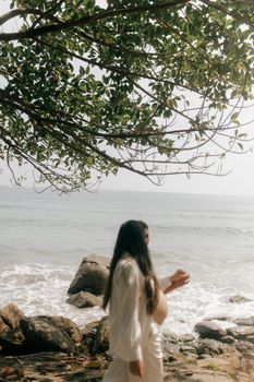 A pregnant woman in white walks along a peaceful rocky beach under leafy branches.